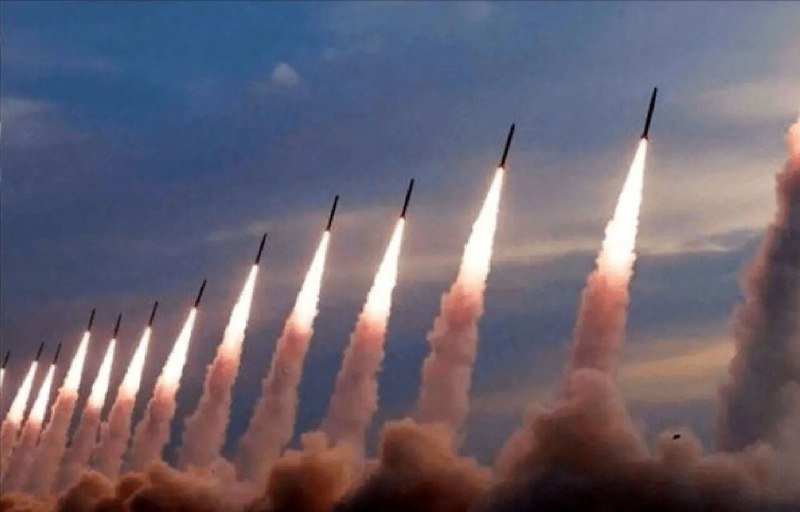 Multiple missiles ascend in a diagonal formation against a partly cloudy sky, each trailing bright flames and thick plumes of smoke.
