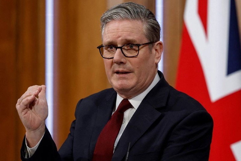 A gray-haired man wearing glasses, a dark suit, and a red tie gestures with a raised fist while speaking, with a Union Jack flag visible in the background.