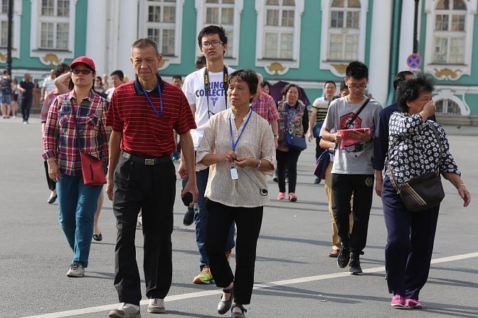 A group of tourists wearing lanyards walks across a paved plaza in front of a green and white ornate building.