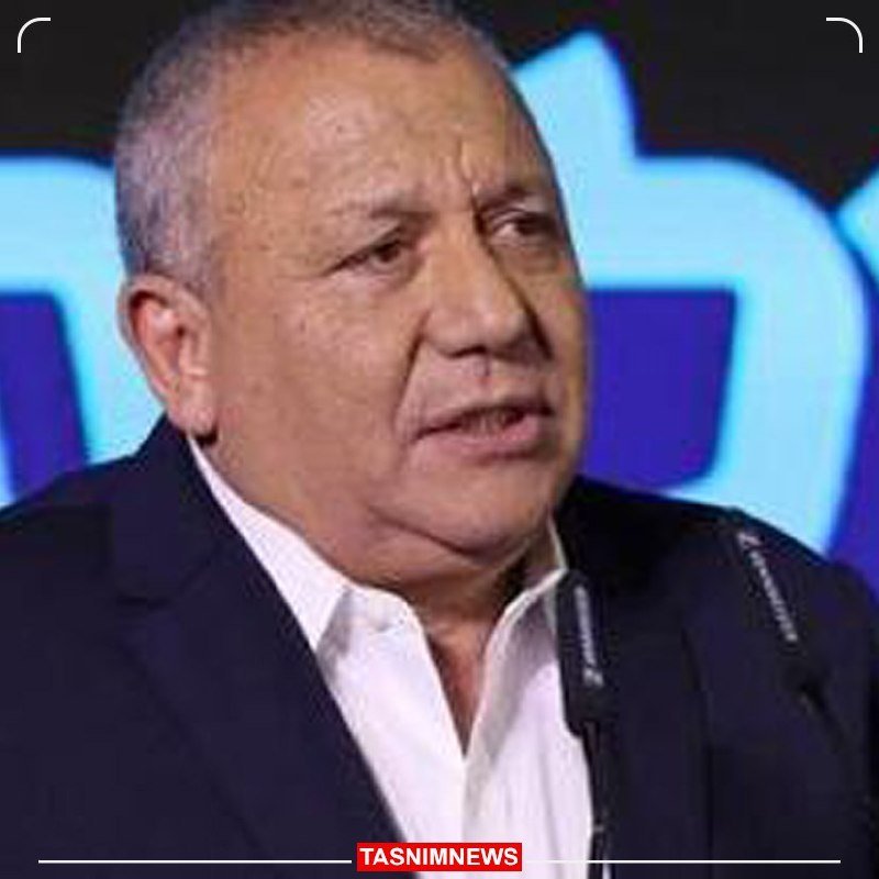An older man in a dark suit and white shirt speaks into a microphone against a blue backdrop, with a "TASNIM NEWS" logo visible at the bottom.