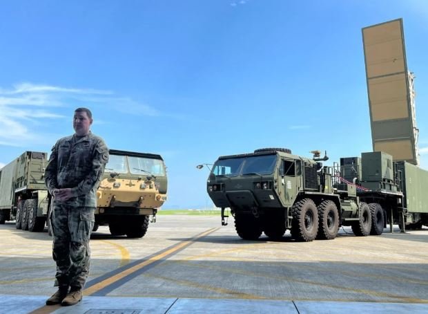 A soldier in camouflage uniform stands on a tarmac beside two large military vehicles, including a truck-mounted radar system with its panel raised vertically.