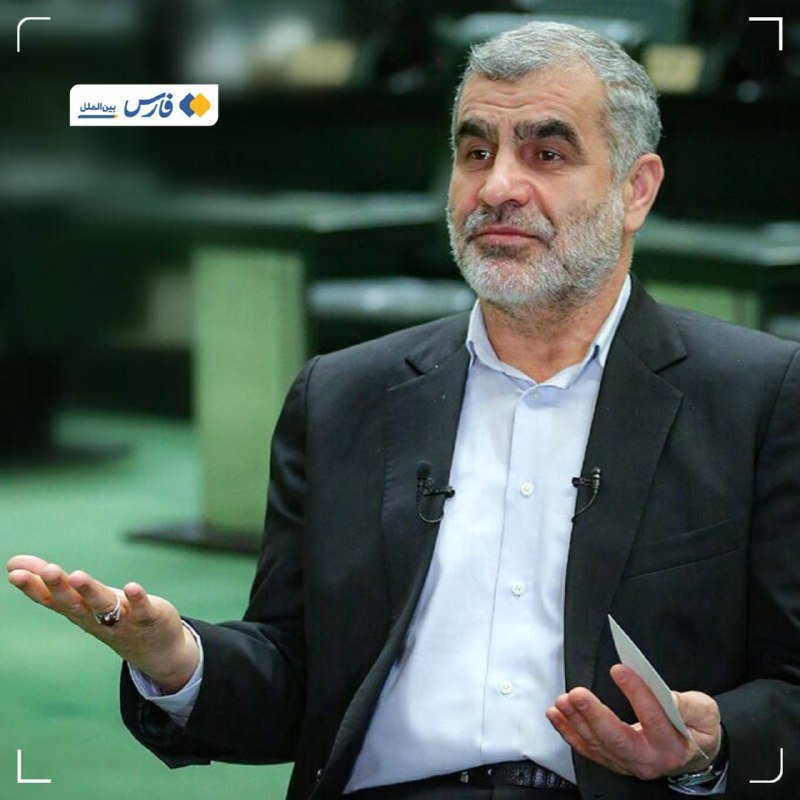 A gray-bearded man in a dark suit and light blue shirt gestures with both hands while holding a small paper, appearing to speak in an indoor setting with green flooring.