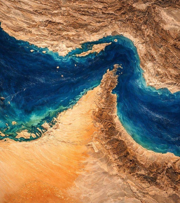 Satellite image showing a narrow landmass separating two bodies of deep blue water, surrounded by arid, orange and brown desert terrain.