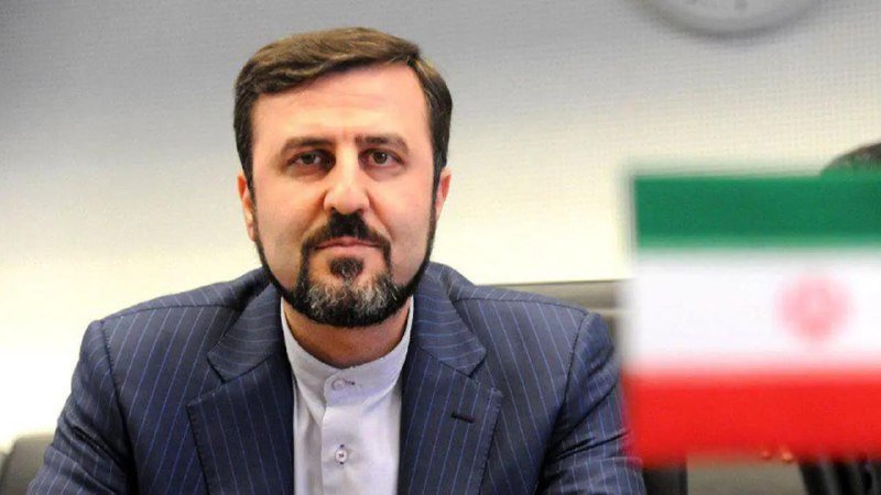 A bearded man in a pinstripe suit and white shirt sits near an Iranian flag, with a partial clock visible on the wall behind him.