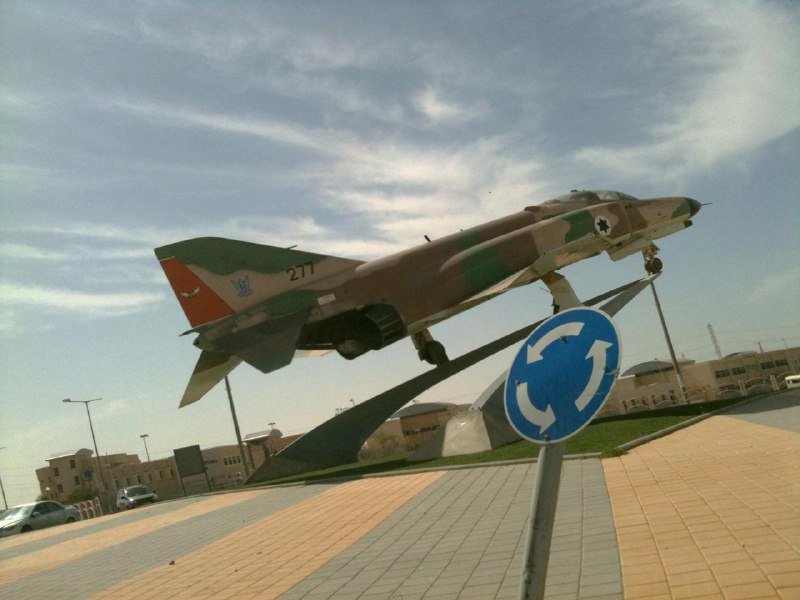 A camouflaged military fighter jet, marked "277," is mounted on a display stand beside a blue roundabout traffic sign, with low buildings and a parked car visible in the background.