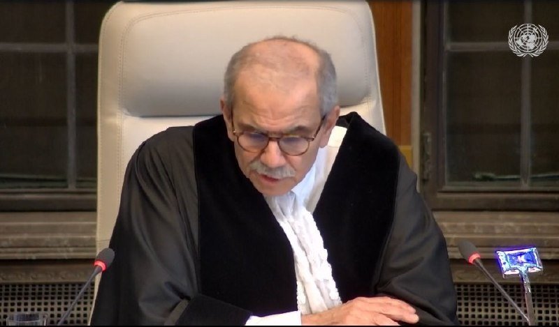 A man in judicial robes and glasses sits in a high-backed white chair, looking downward, with a United Nations emblem visible on the wall behind him.