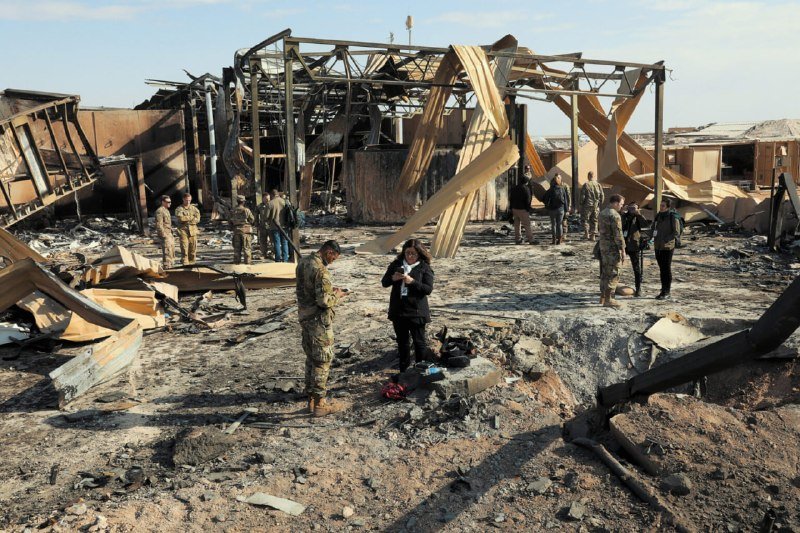 Soldiers and civilians stand among the wreckage of a destroyed building with twisted metal beams and scattered debris under a clear sky.