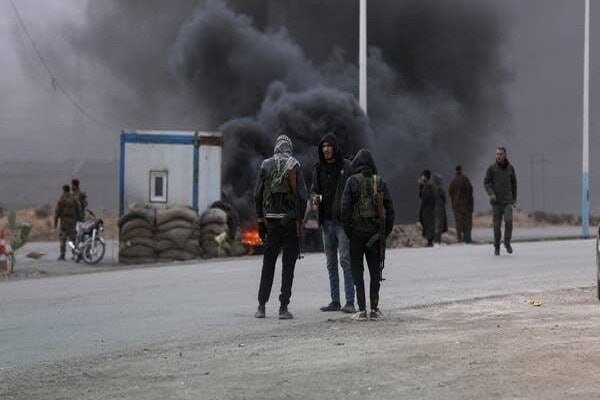 Masked men stand on a road as a fire burns behind them, sending thick black smoke into the sky.