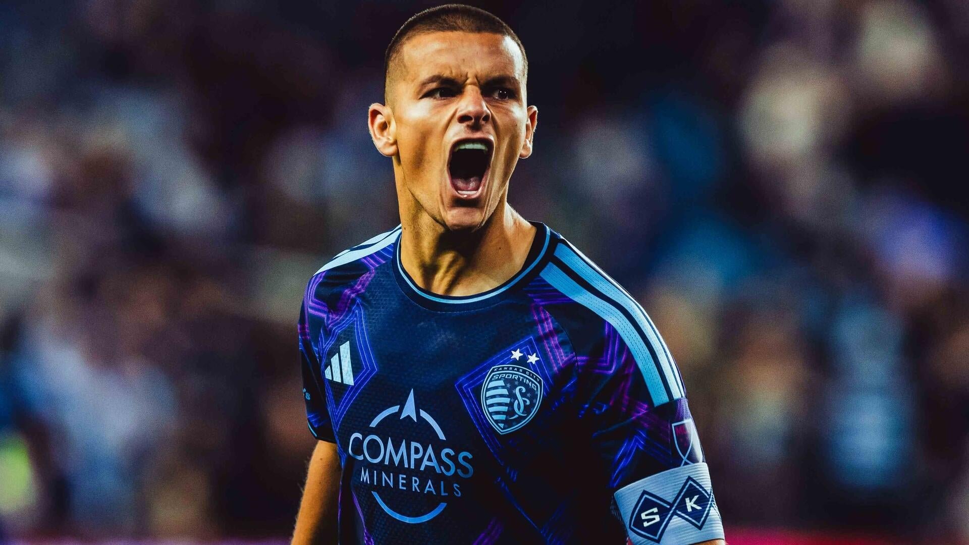 A soccer player wearing a dark Sporting Kansas City jersey with "Compass Minerals" sponsorship shouts intensely during a match, with a blurred crowd in the background.
