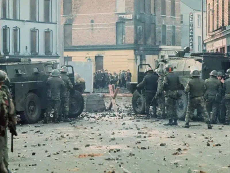 Soldiers in helmets and camouflage uniforms stand near armored vehicles on a debris-littered street in front of brick buildings, including one with a "CENTRAL CAFE" sign.
