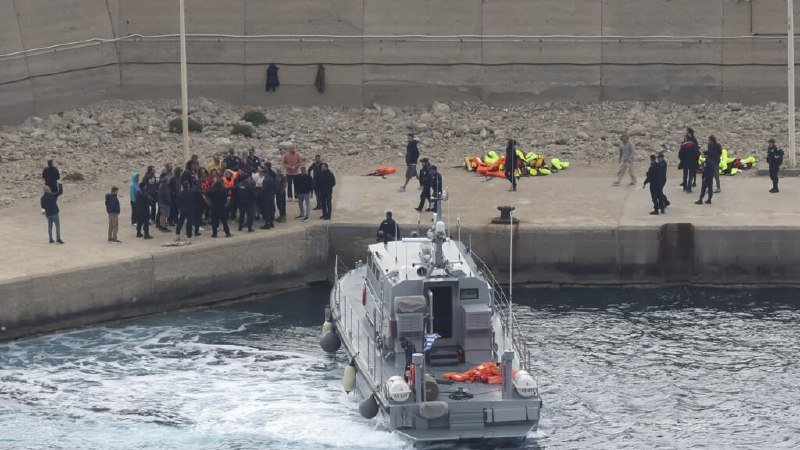 A gray patrol boat maneuvers in a harbor alongside a concrete pier, where uniformed personnel, civilians, and orange life jackets are gathered on the rocky shore.