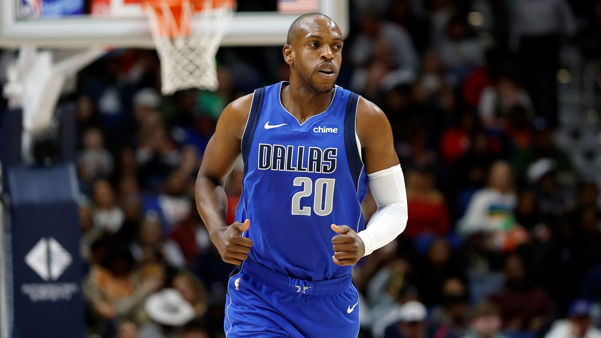 A Dallas Mavericks player wearing jersey number 20 runs on the court during an NBA game, with a basketball hoop and crowd visible behind him.