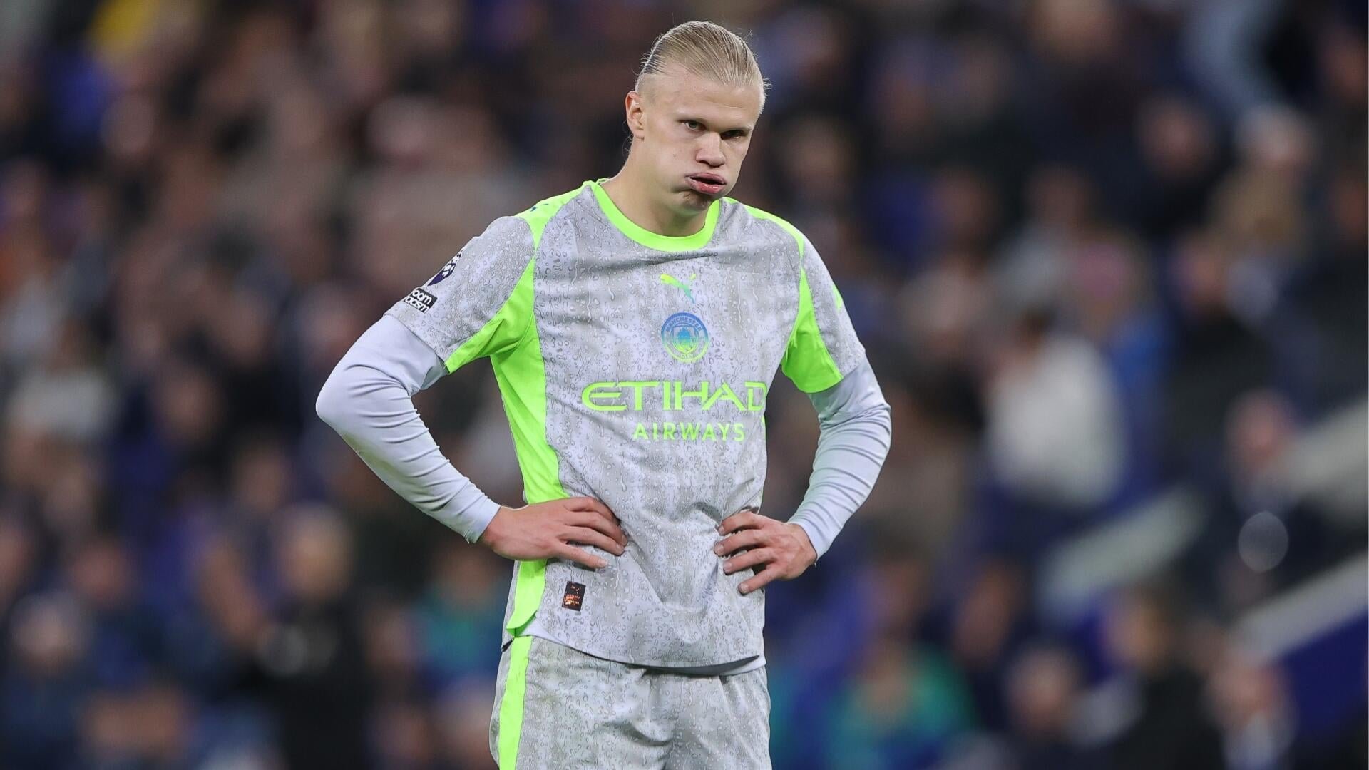A blonde soccer player in a grey Manchester City jersey stands with hands on hips, sticking his tongue out during a match.
