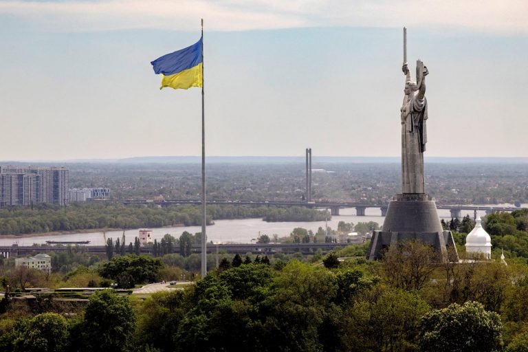 A large blue-and-yellow flag flies on a tall pole beside a towering metal statue of a woman holding a sword aloft, overlooking a river, bridge, and cityscape.