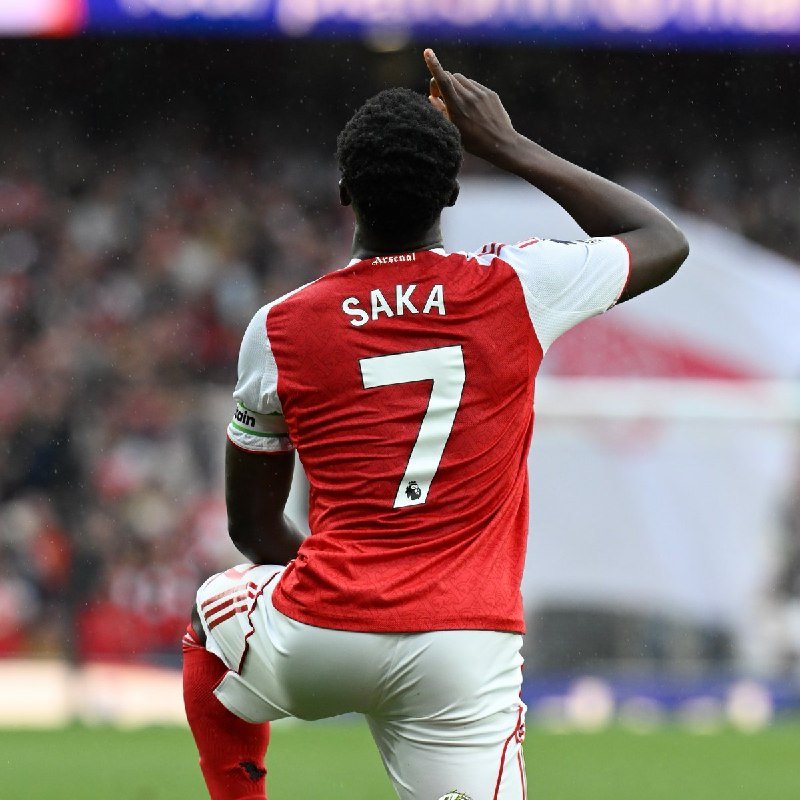 A soccer player wearing a red and white jersey with "SAKA" and the number 7 on the back raises his right index finger while kneeling on a stadium field.