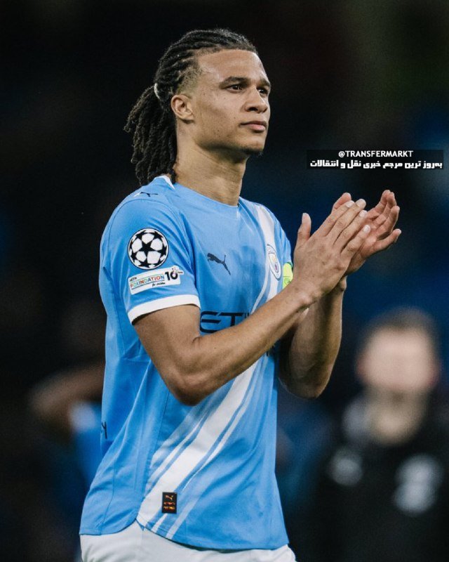 A soccer player with long braided hair, wearing a light blue jersey with a Champions League patch, claps his hands while looking forward on the field.