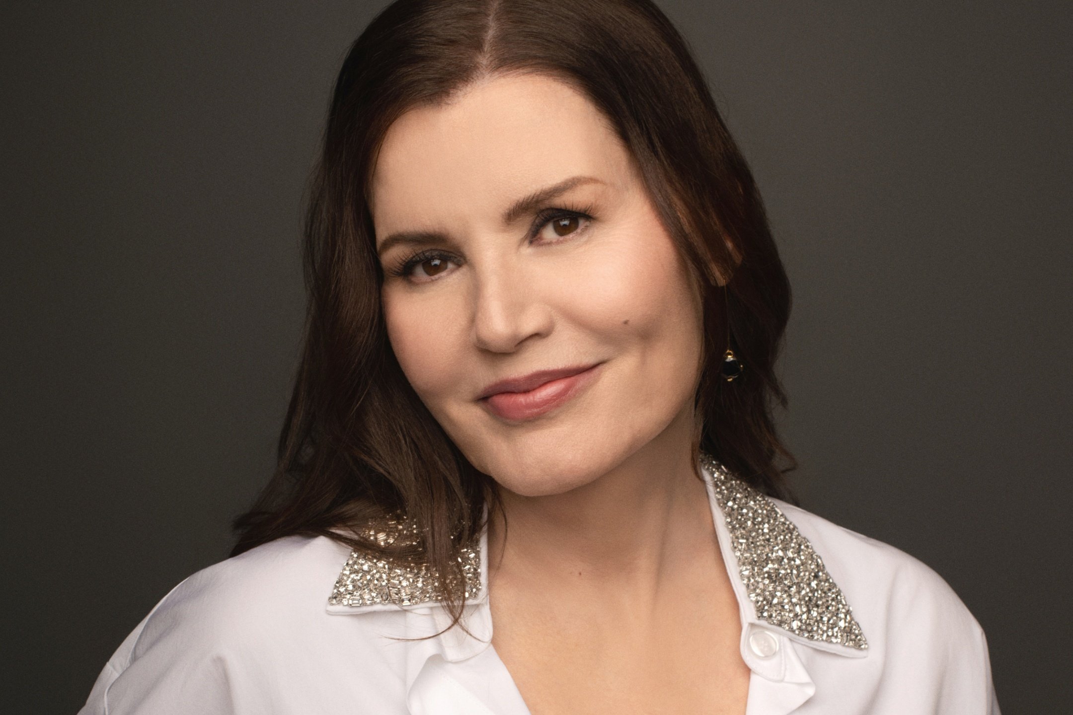 A woman with long dark hair poses for a portrait against a gray background, wearing a white button-up shirt with rhinestone-embellished collars.