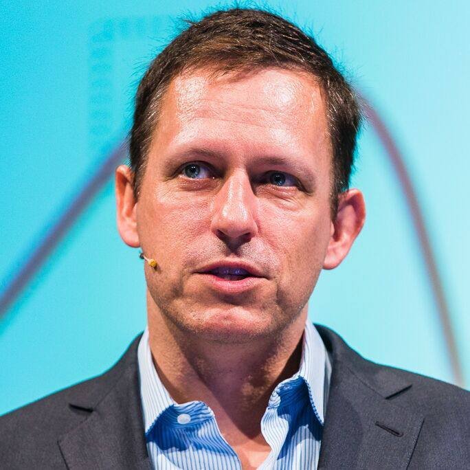 A man wearing a dark suit jacket and light blue shirt speaks with a small earpiece microphone against a teal background.