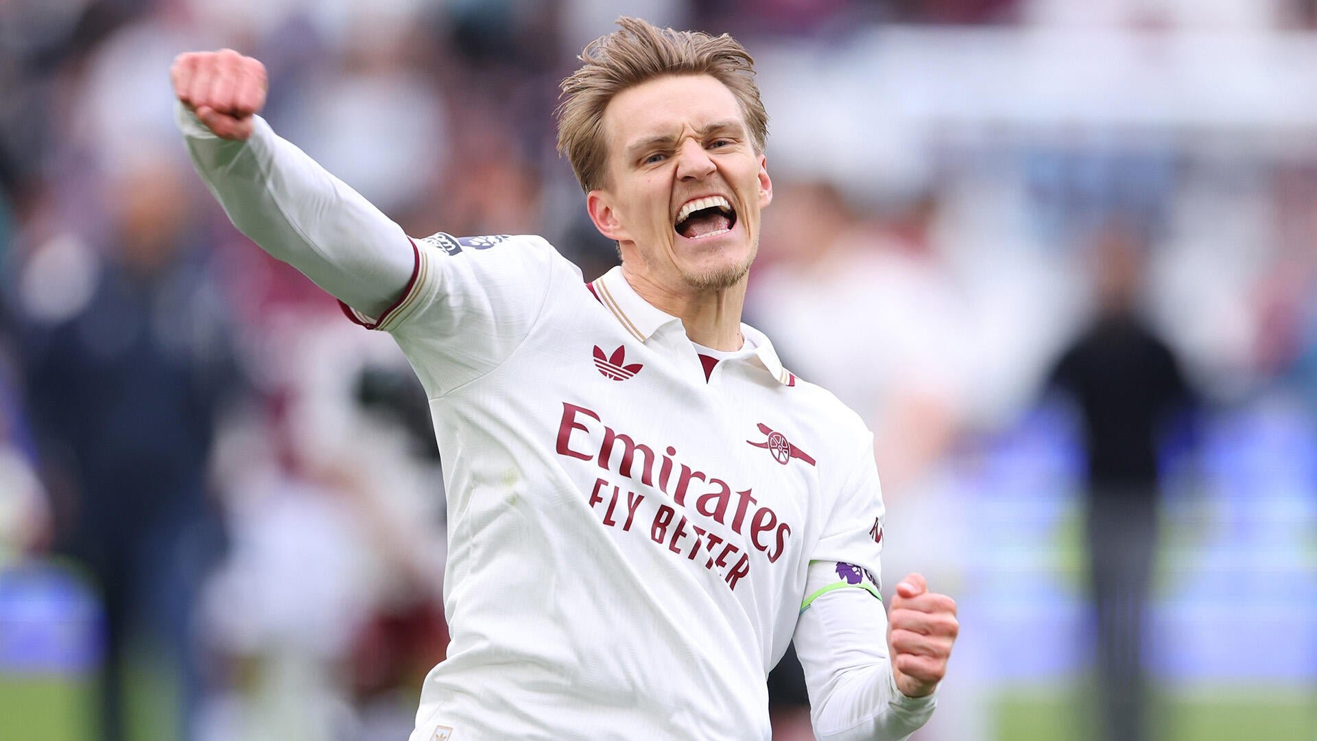 A blonde soccer player in a white Arsenal jersey with "Emirates Fly Better" sponsorship celebrates with a raised fist and open-mouthed shout before a blurred stadium crowd.