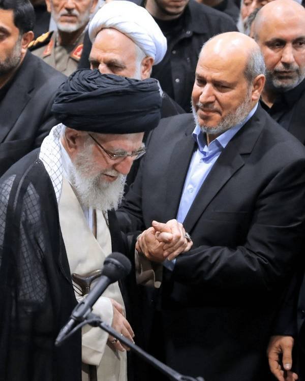 A bearded cleric in black turban and robes shakes hands with a suited, bearded man before a microphone, surrounded by a crowd of men.