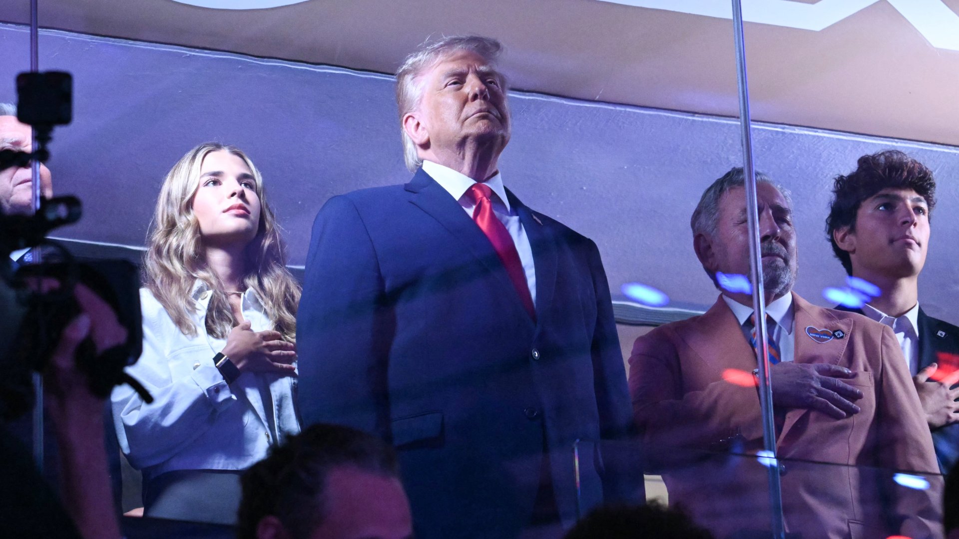 A man in a blue suit and red tie stands solemnly alongside three others with hands over their hearts in a dimly lit venue.