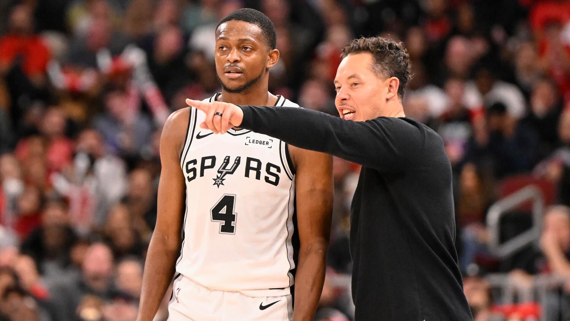 A man in a black shirt points and smiles while speaking to a basketball player wearing a white Spurs jersey with the number 4 during a game in front of a crowd.