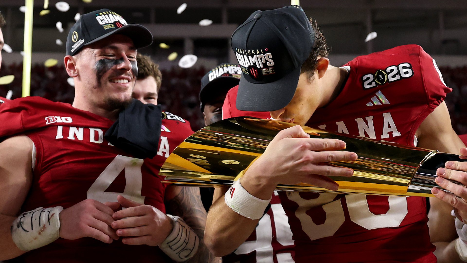 Indiana football players in red jerseys and "National Champs" hats embrace and kiss a gold trophy amid falling confetti.