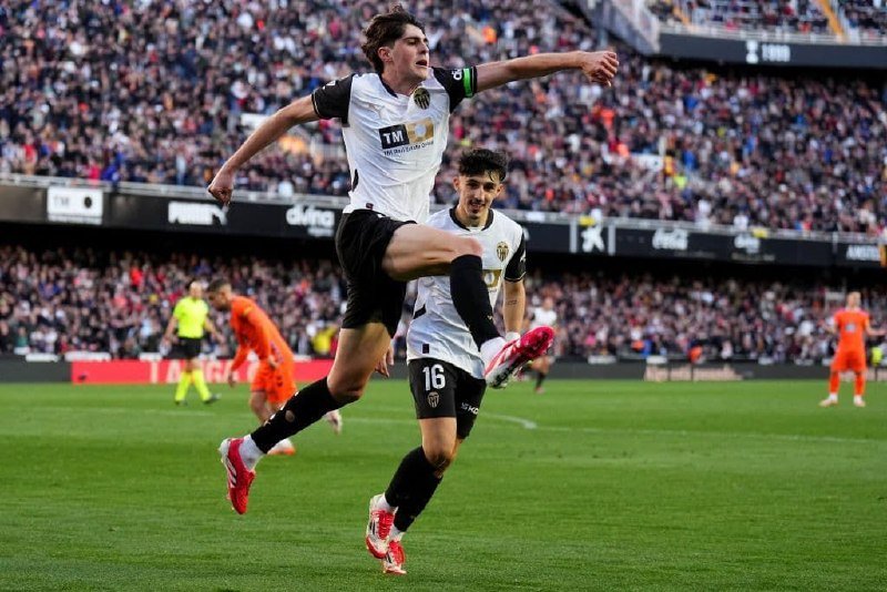Two soccer players in white and black uniforms celebrate on the pitch, one leaping mid-air, while a player in orange and a referee appear in the background before a crowded stadium.