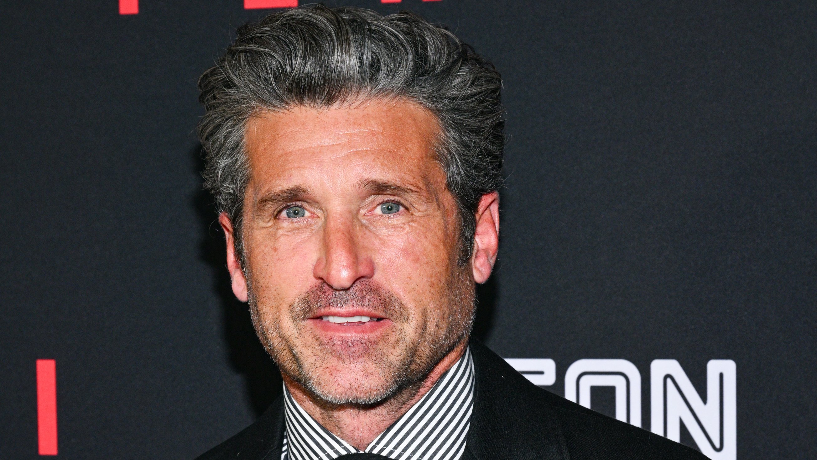 A man with gray-streaked hair and stubble wears a black suit jacket and striped shirt at an event.