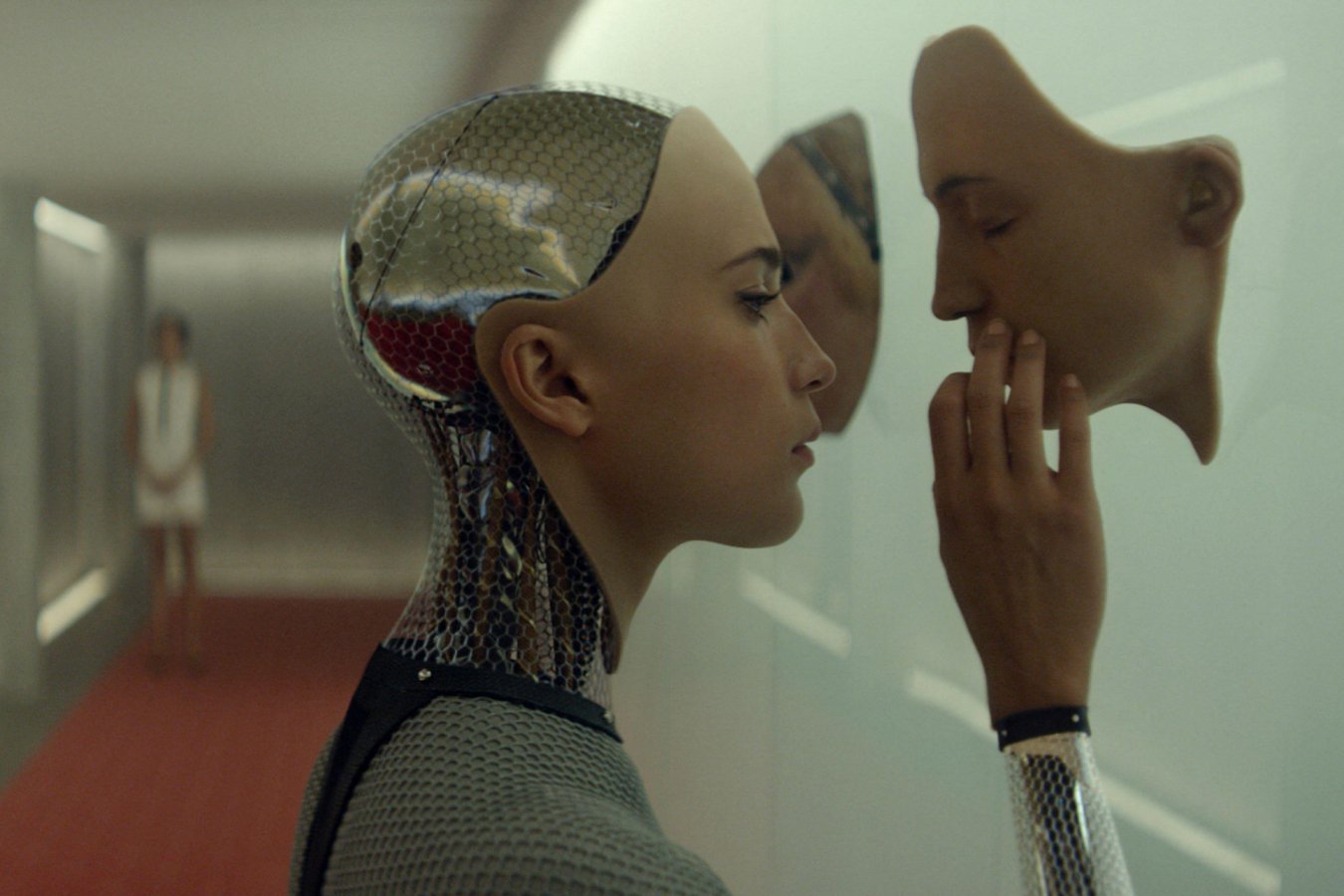 A humanoid android with a metallic mesh skull holds a flesh-toned face mask up to its own face in a sterile white corridor, while another figure stands in the background.
