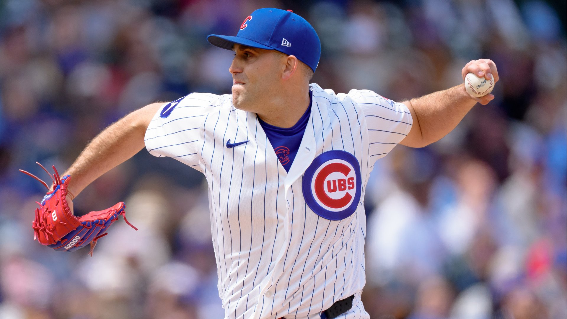 Chicago Cubs pitcher winds up to throw a baseball during a daytime game, wearing a white pinstriped jersey and blue cap with a blurred crowd in the background.