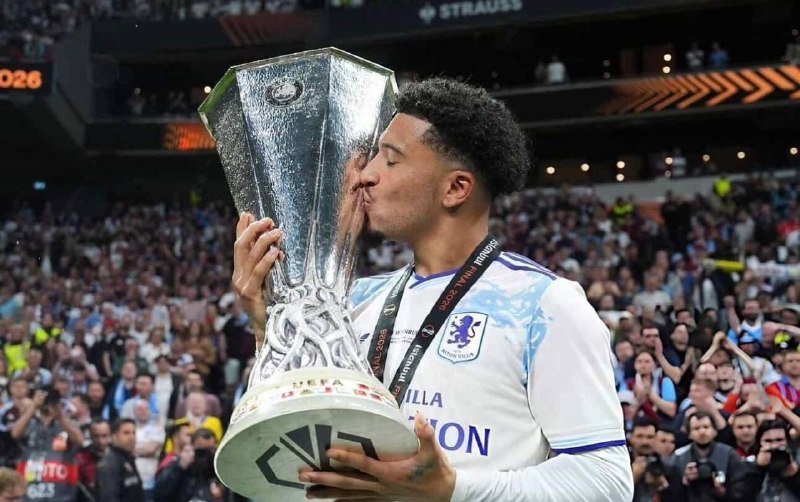 A soccer player in a light blue and white Aston Villa jersey kisses a large silver trophy in a crowded stadium.