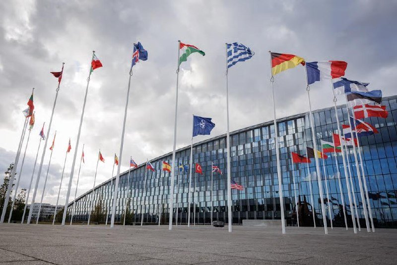 Multiple national flags fly on tall poles in front of a large glass-fronted building under a cloudy sky.