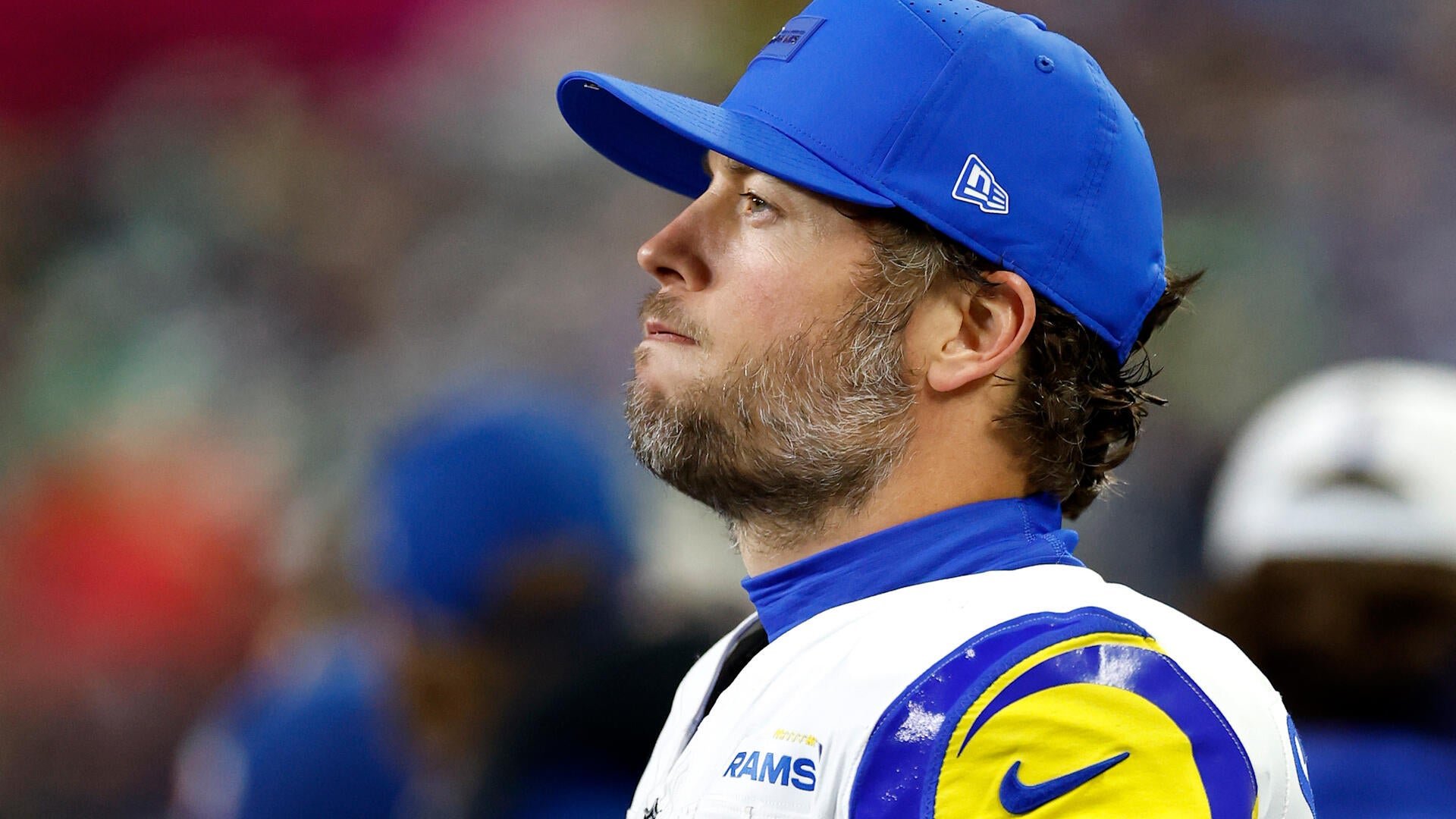 A bearded man in a blue cap and white Rams jersey looks off to the side during a football game, with a blurred crowd in the background.