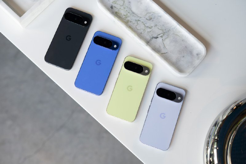 Four smartphones in black, blue, green, and light purple, each marked with a "G" logo, rest on a white surface beside a marble tray.