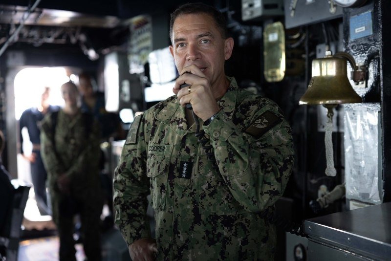 A U.S. Navy officer in camouflage uniform speaks into a microphone aboard a ship, with a brass bell mounted on a wall panel behind him and other personnel standing in the background.