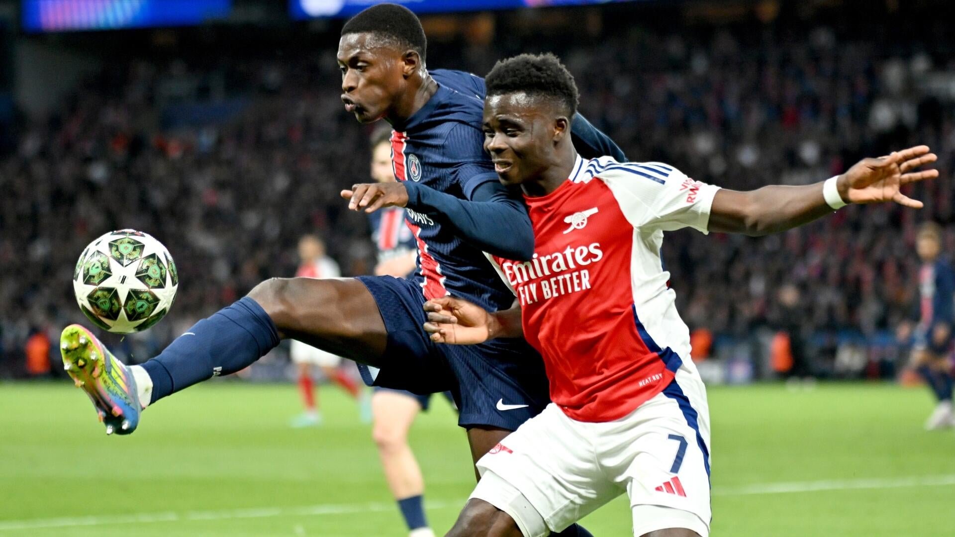 Two soccer players, one in a dark blue kit and one in a red and white Arsenal jersey, compete for the ball mid-air during a match.