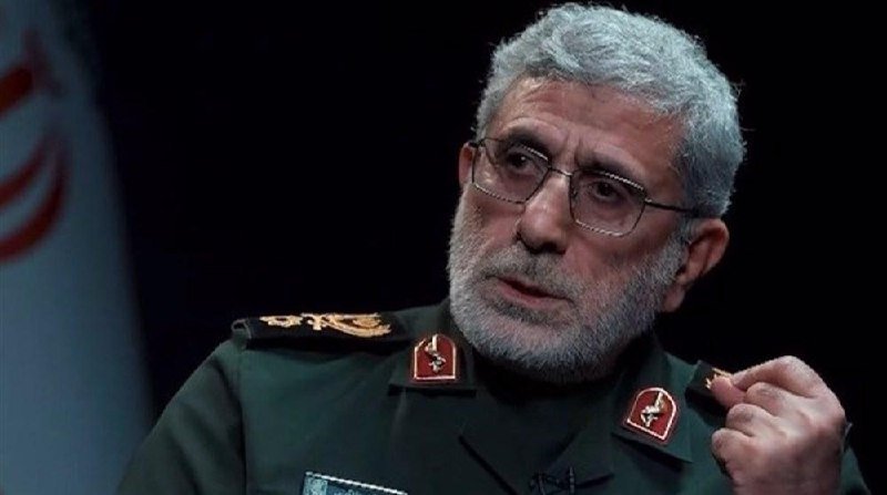 A gray-bearded man in a green military uniform with insignia and rank markings speaks while gesturing, seated in front of a dark background with a partially visible flag.