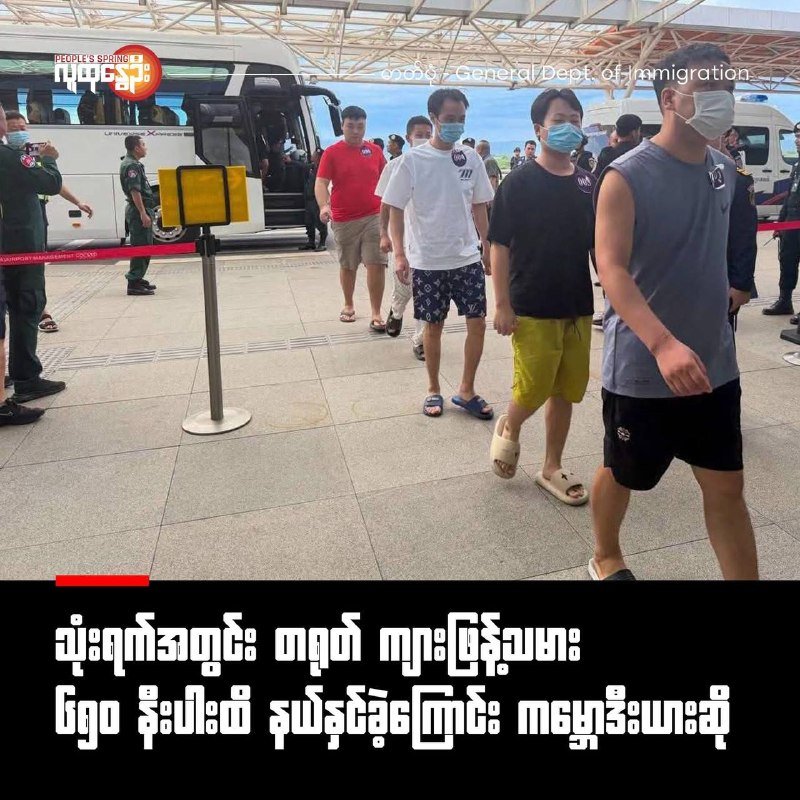 Masked men in casual clothing walk in a line past uniformed personnel and buses on a tiled terminal platform.