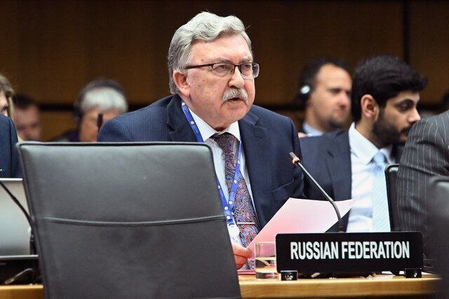 A gray-haired, mustached man in a suit and glasses holds papers beside a "RUSSIAN FEDERATION" placard at a formal conference table with other attendees.