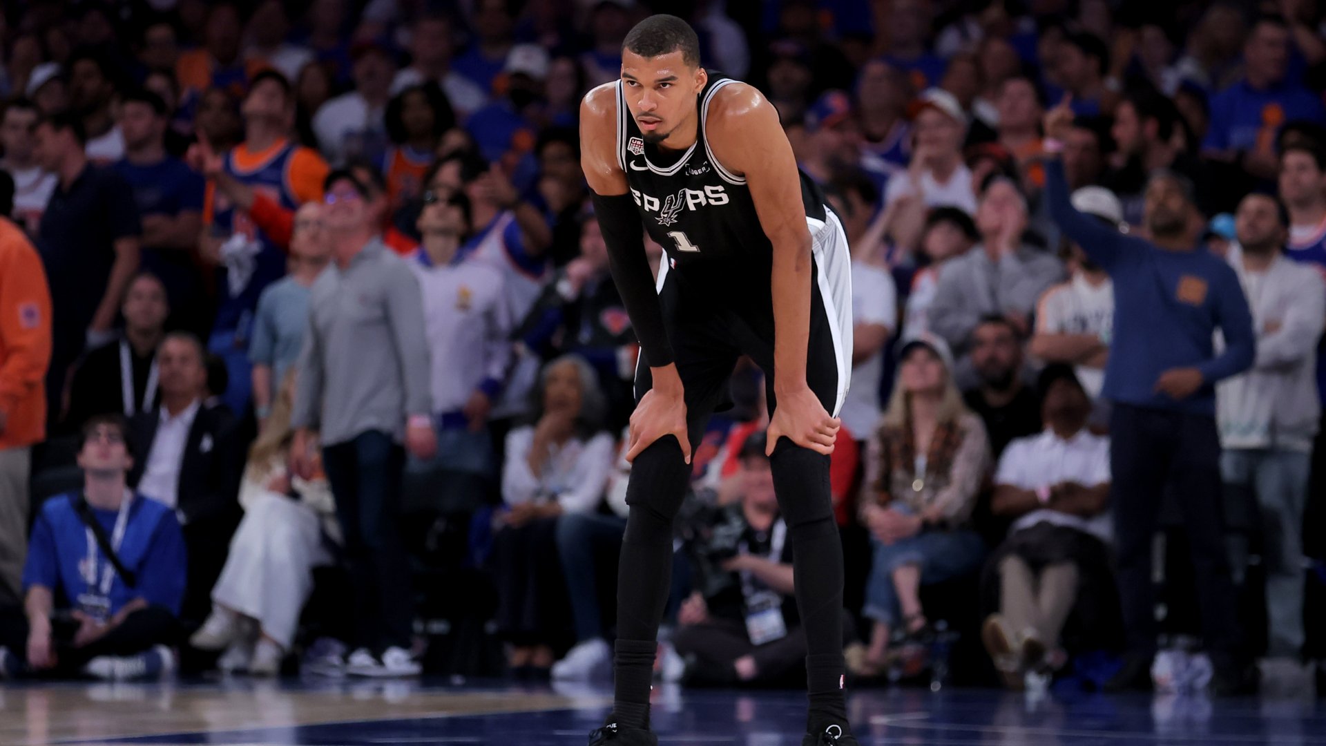 A basketball player in a black San Antonio Spurs jersey stands bent over with hands on his knees, facing a crowded arena of spectators during a game.