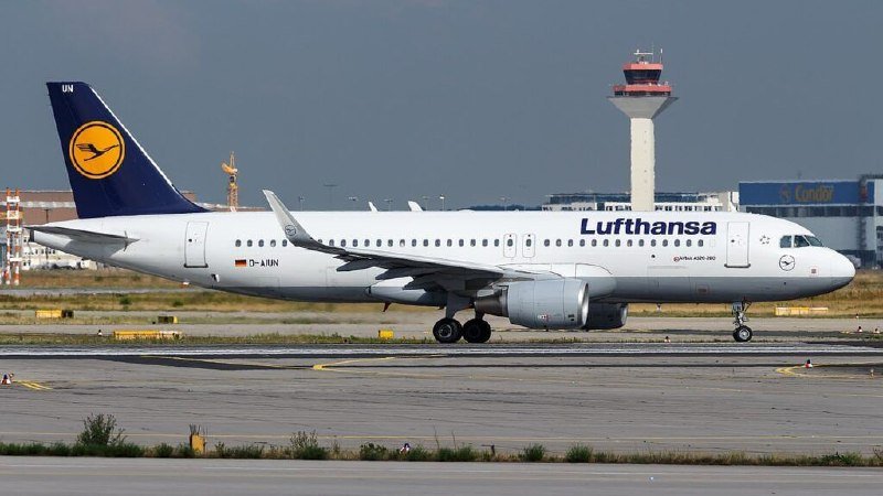 A Lufthansa passenger jet taxis on a runway, with an air traffic control tower and airport buildings visible in the background.