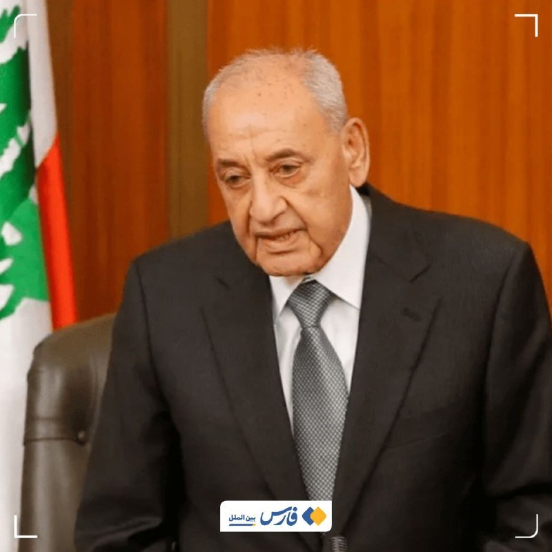 An elderly man in a dark suit and gray tie sits beside a Lebanese flag, looking downward in a formal setting with orange curtains.