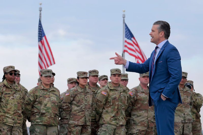 A man in a blue suit points toward a formation of soldiers in camouflage uniforms, with two American flags visible in the background.