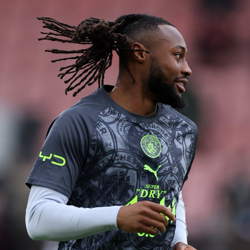 A soccer player with long dreadlocks wearing a dark Manchester City training shirt with neon green Puma and Super Dry logos warms up on a blurred pink-toned background.