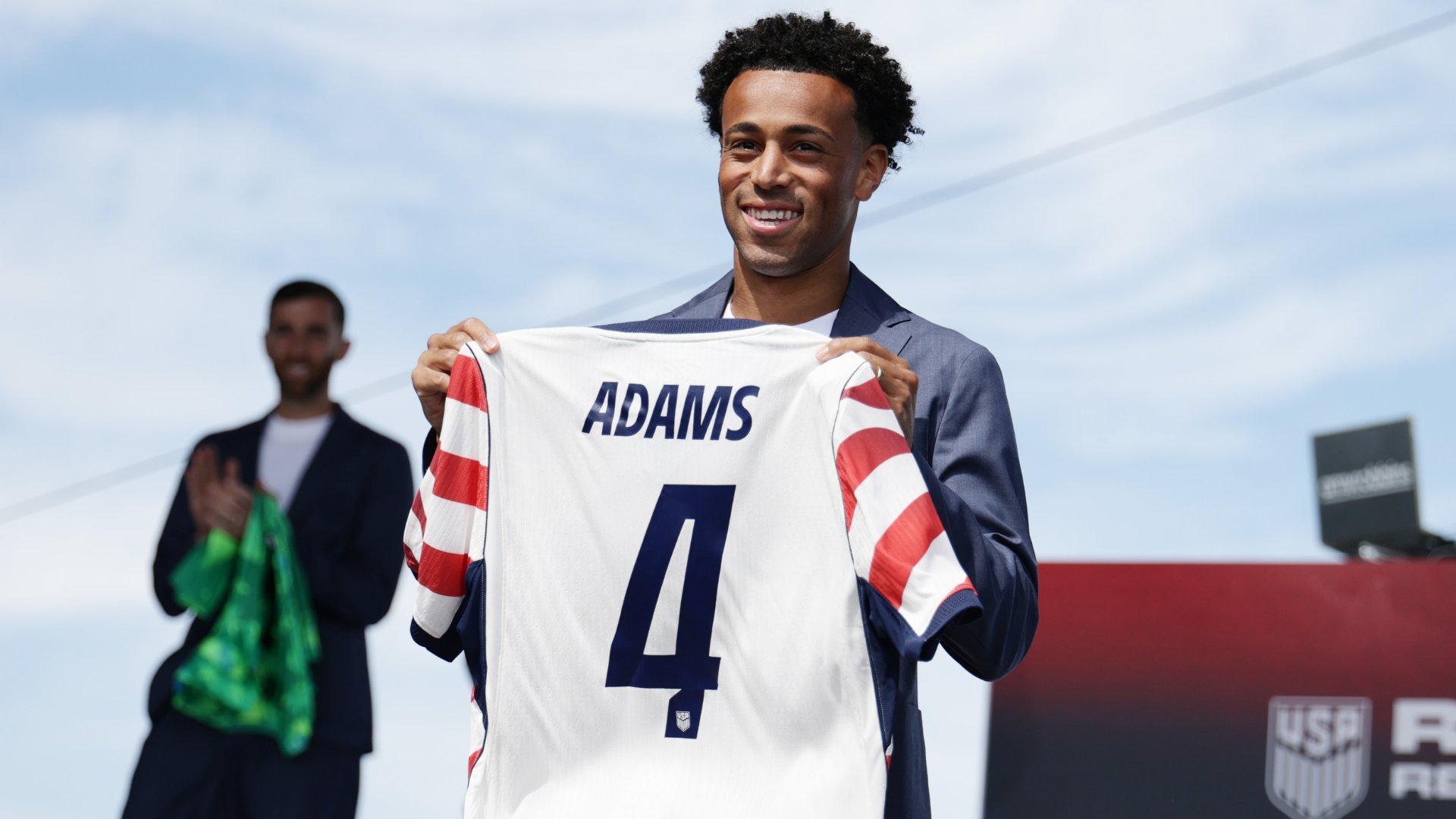 A smiling man in a navy suit holds up a white "ADAMS" #4 USA jersey, while another man applauds in the background.
