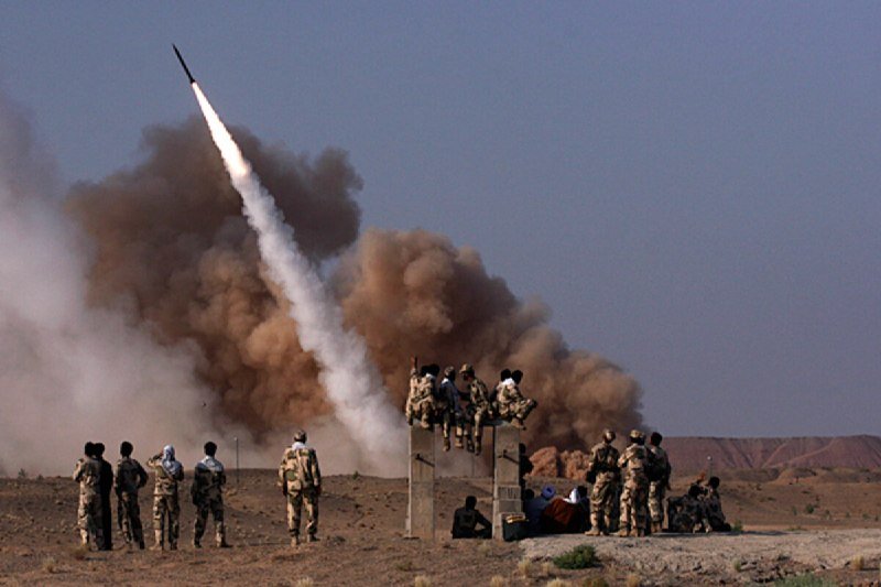Soldiers in desert camouflage watch as a missile launches skyward, leaving a bright trail and a large cloud of brown dust and smoke behind it.