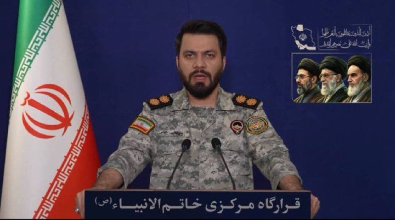 A bearded man in a camouflage military uniform stands at a podium with microphones, beside an Iranian flag, with portraits of three clerics displayed on the wall behind him.