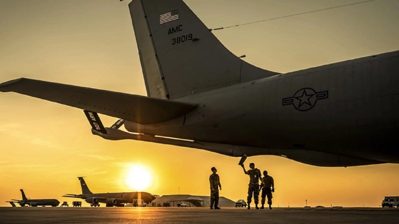 Silhouetted personnel conduct maintenance beneath the tail section of a large military aircraft marked "AMC 38019" against a sunset-lit tarmac with additional aircraft visible.