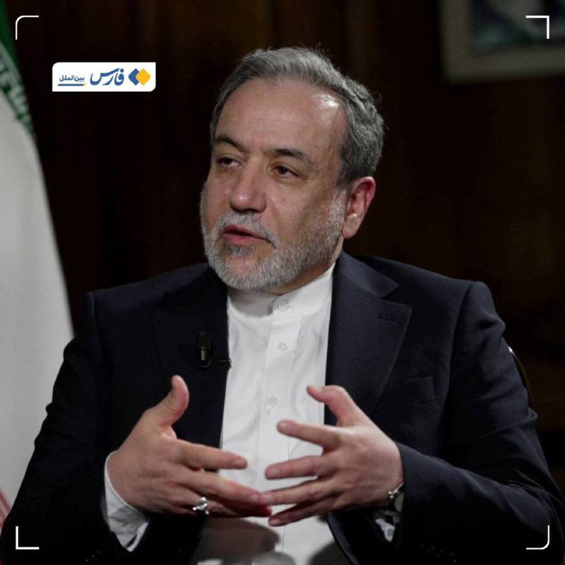 A gray-bearded man in a dark suit and white shirt gestures with both hands while speaking during an interview, with an Iranian flag visible behind him.